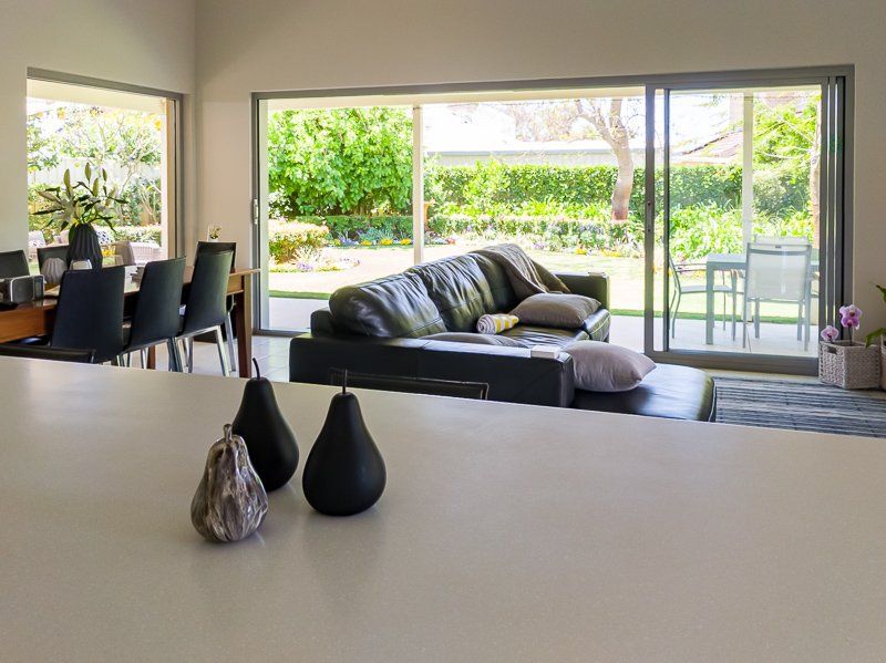 Modern living room with open windows, leather sofa, dining table, and outdoor garden view.