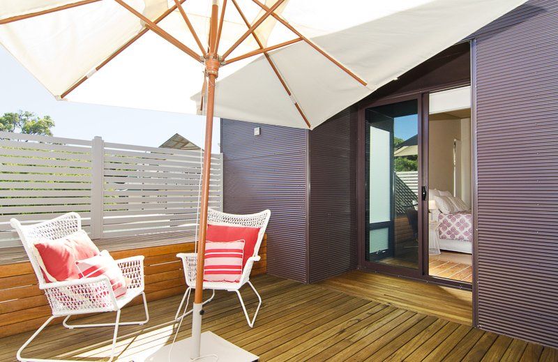 Wooden deck with white chairs, red pillows, and umbrella in front of a building with sliding glass doors.