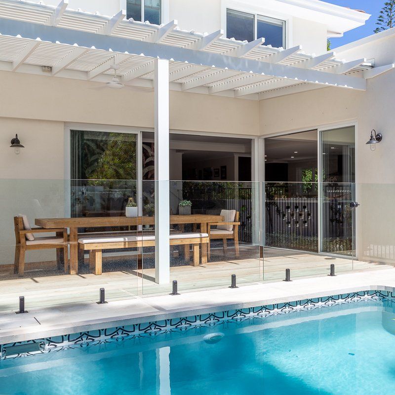 Poolside patio with wooden table, seating, and pergola. Glass fence surrounds the pool.