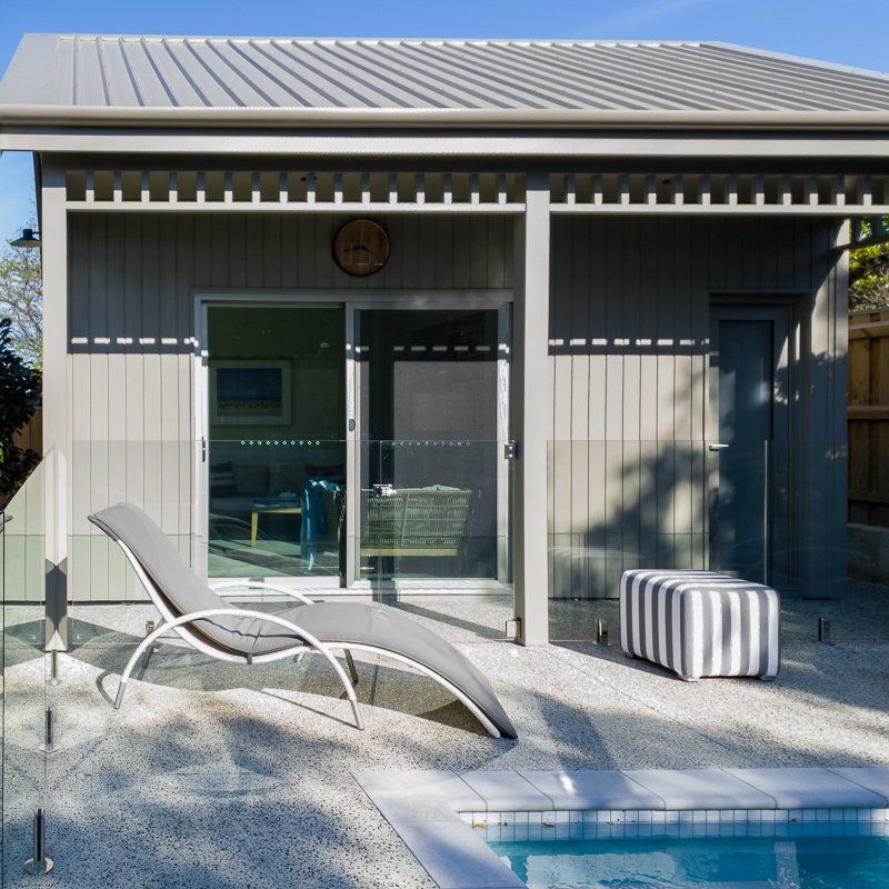 Outdoor relaxation area by a pool; grey building, lounge chair, striped ottoman, speckled ground.