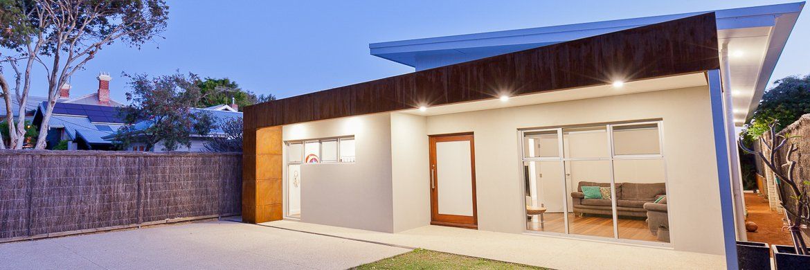 Modern building with a brown roof and white walls. Includes a wooden door and large windows. Dusk setting.