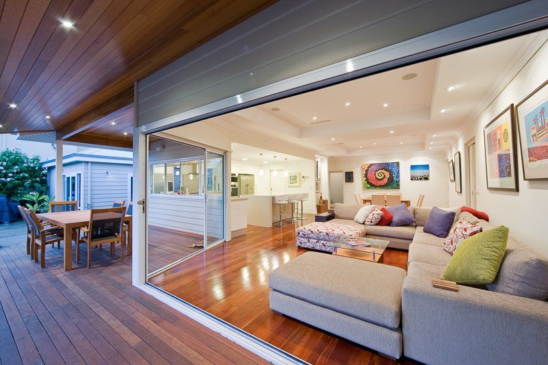 Open-plan living space with outdoor deck; glass doors; wooden flooring; sectional sofa; kitchen visible in the background.