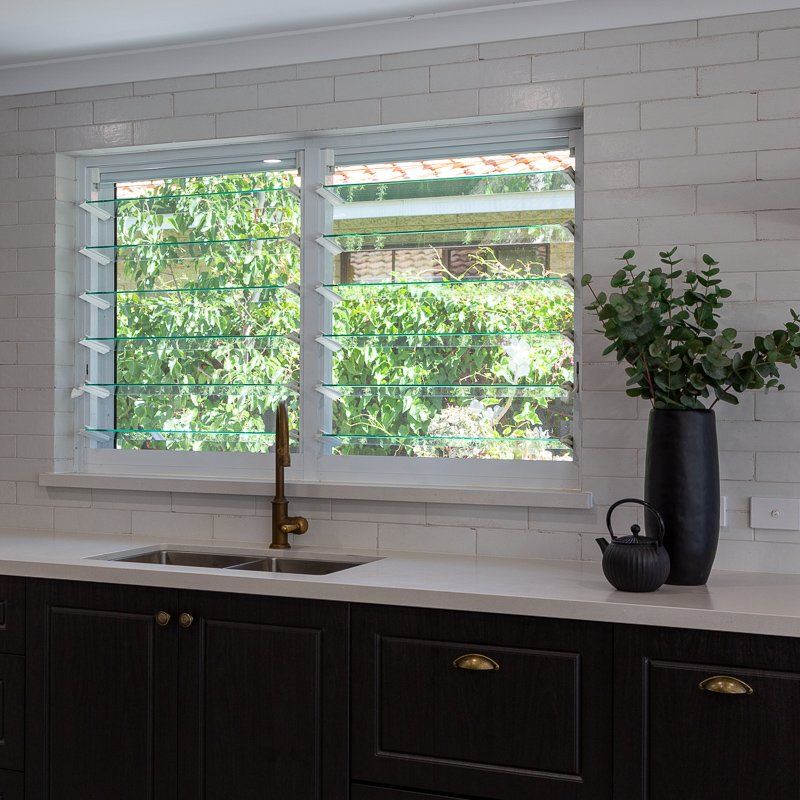 Kitchen with a white countertop, dark cabinets, a sink, and a window with green-tinted louvers overlooking greenery.