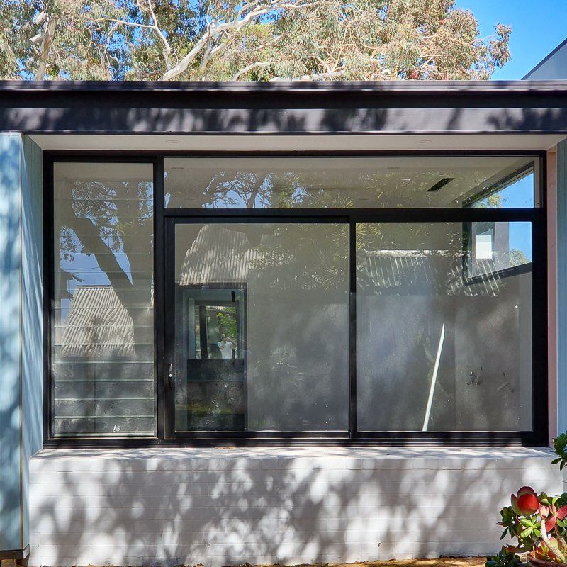 Black-framed window on a light blue building, reflecting trees and a doorway. Sunny day.