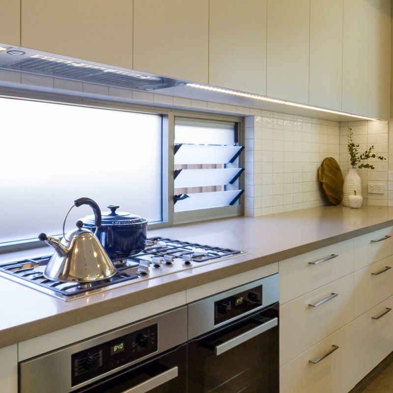 Modern kitchen with stovetop, ovens, and cabinets; window above with a kettle and pot.