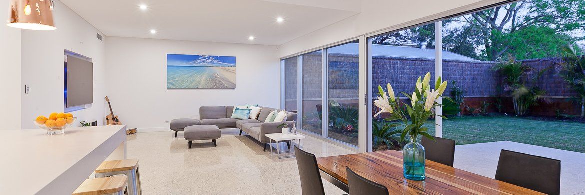 Open-plan living room with white walls, gray sofa, beach artwork, dining table, and view to a backyard.