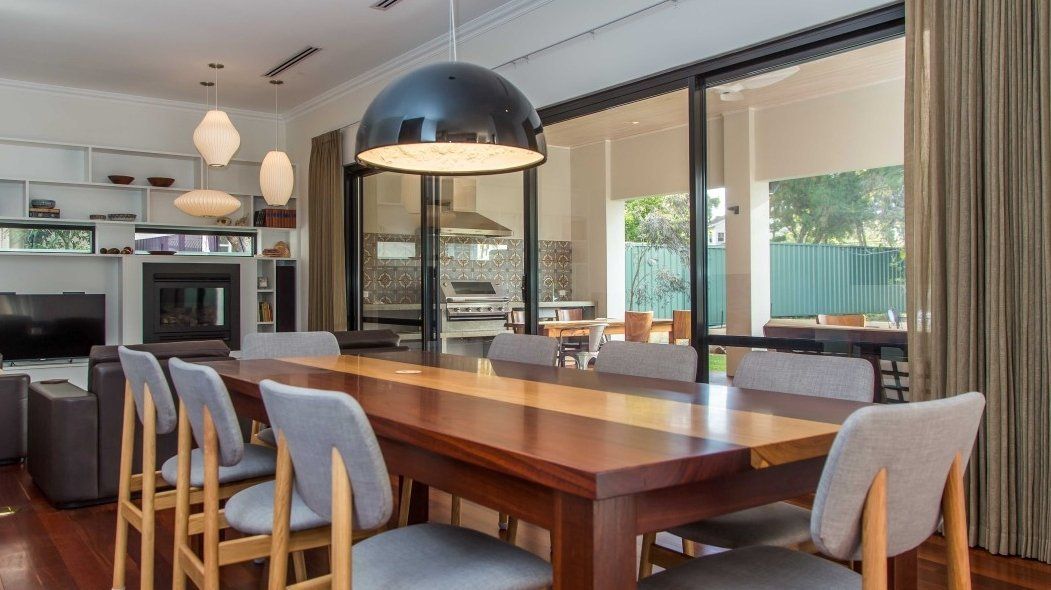 Dining room with a long wooden table and chairs, connected to an outdoor kitchen through glass doors.