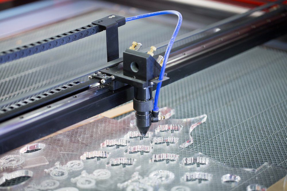 Laser cutter in action, cutting shapes from clear acrylic sheet. Blue air hose and black machine.