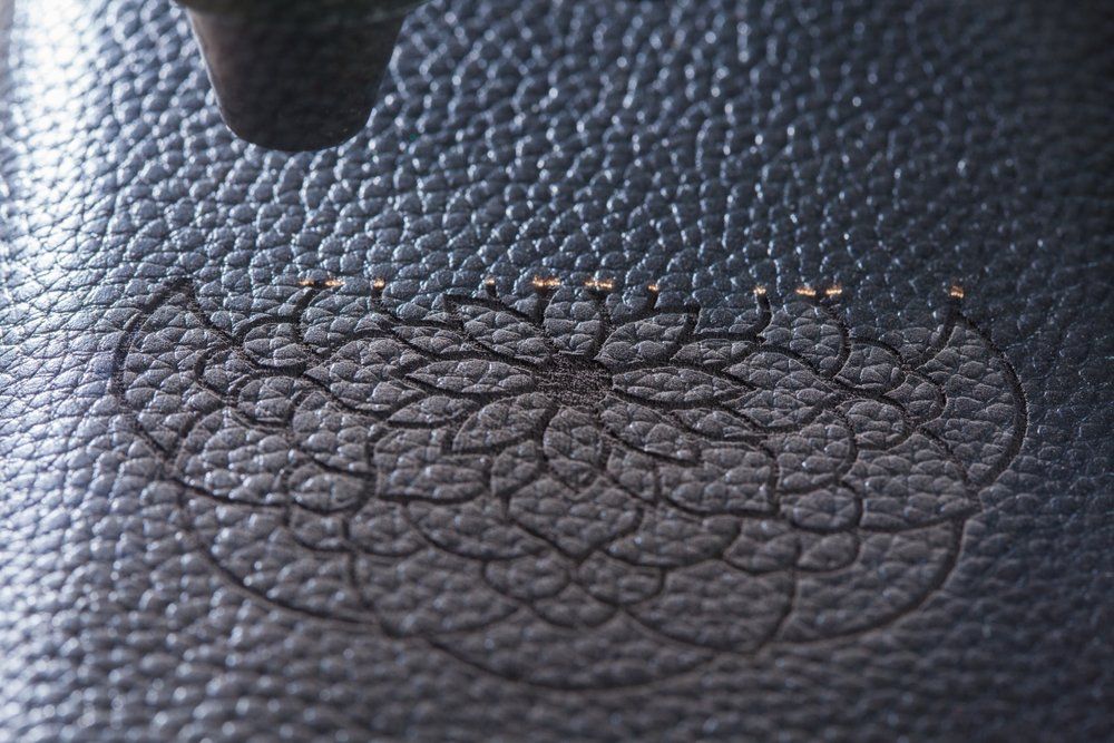 Laser etching floral design on dark leather. A laser engraver is visible above the design.