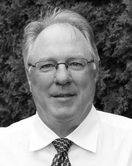 Man in glasses and tie, smiling, against a blurred outdoor background.