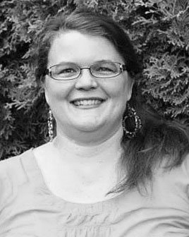 Woman with glasses smiles, wearing earrings and a light shirt, set against leafy green background.