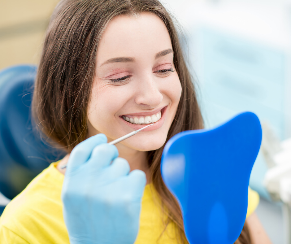 Woman in yellow shirt smiles, examines teeth with a mirror and dental tool. Blue gloves and a dental chair.