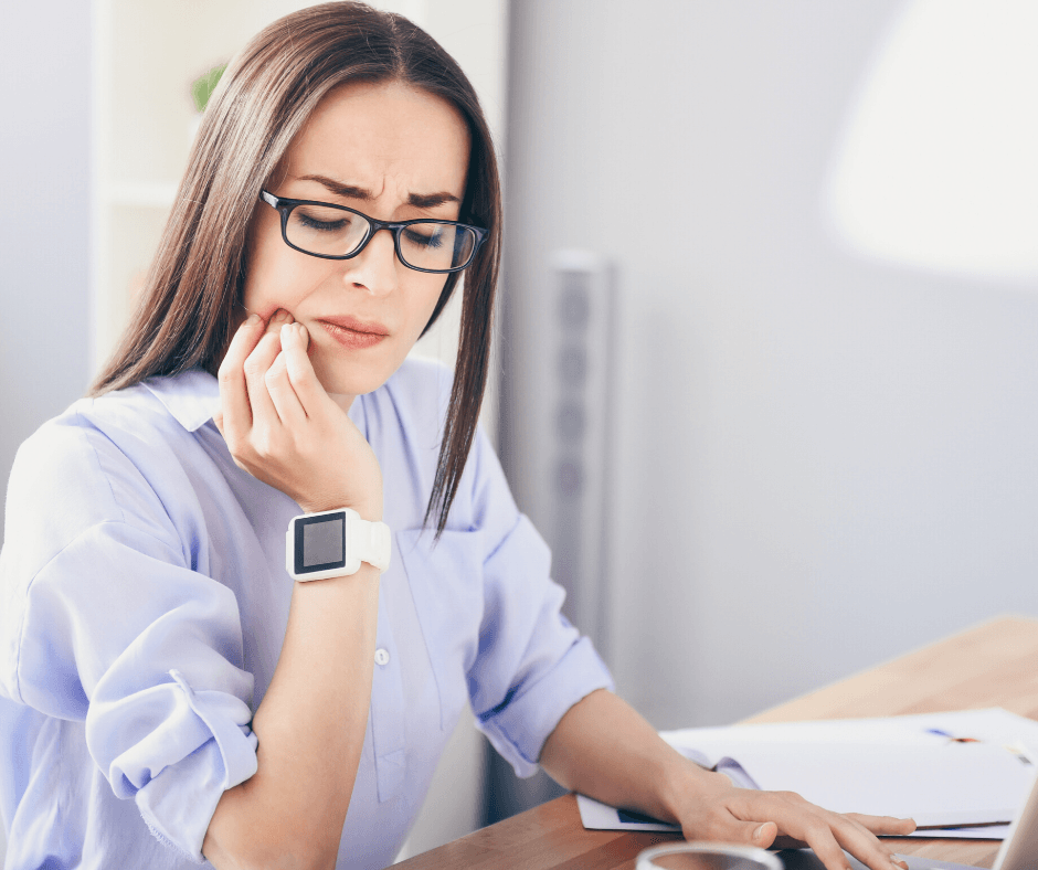 Woman with glasses holding her jaw, appearing to be in pain while working at a desk.