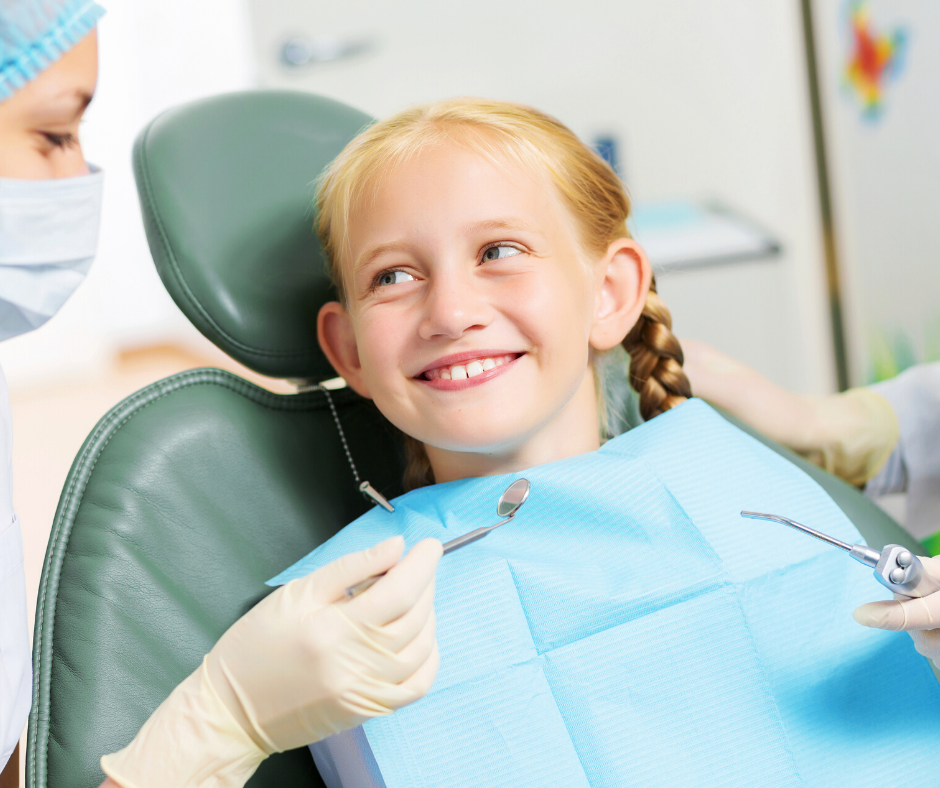 Girl in dental chair smiles at dentist with tools. Dentist wears a mask and gloves.