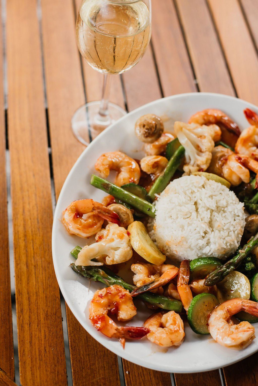 A plate of food with shrimp , rice and vegetables next to a glass of wine on a wooden table.