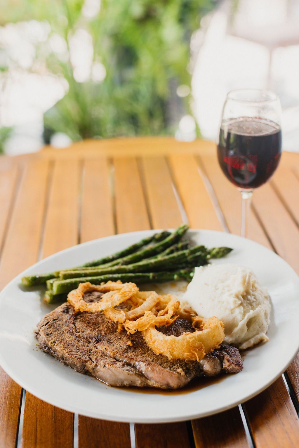 A white plate topped with steak , mashed potatoes , asparagus and onion rings next to a glass of wine