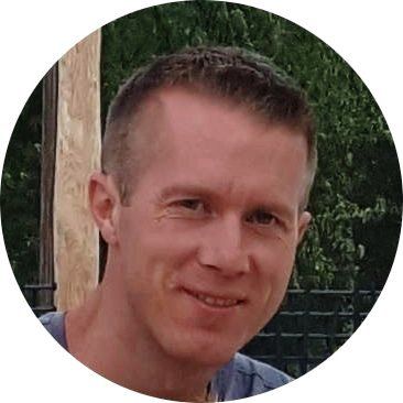 Een man met kort bruin haar glimlacht, buiten bij een houten paal en groen.