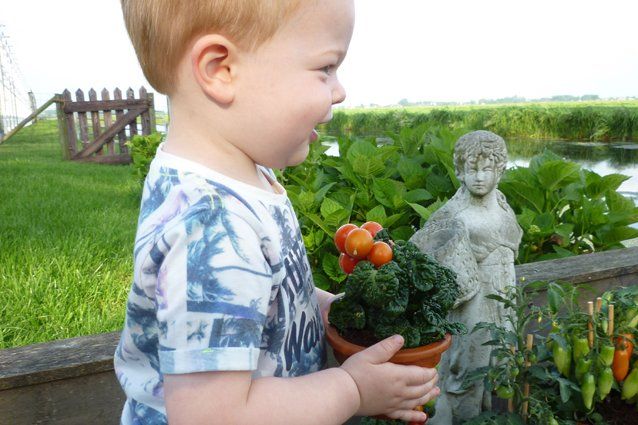 Jong kind met een potplant in zijn handen, lachend. Buiten, standbeeld, tuin, water op de achtergrond.