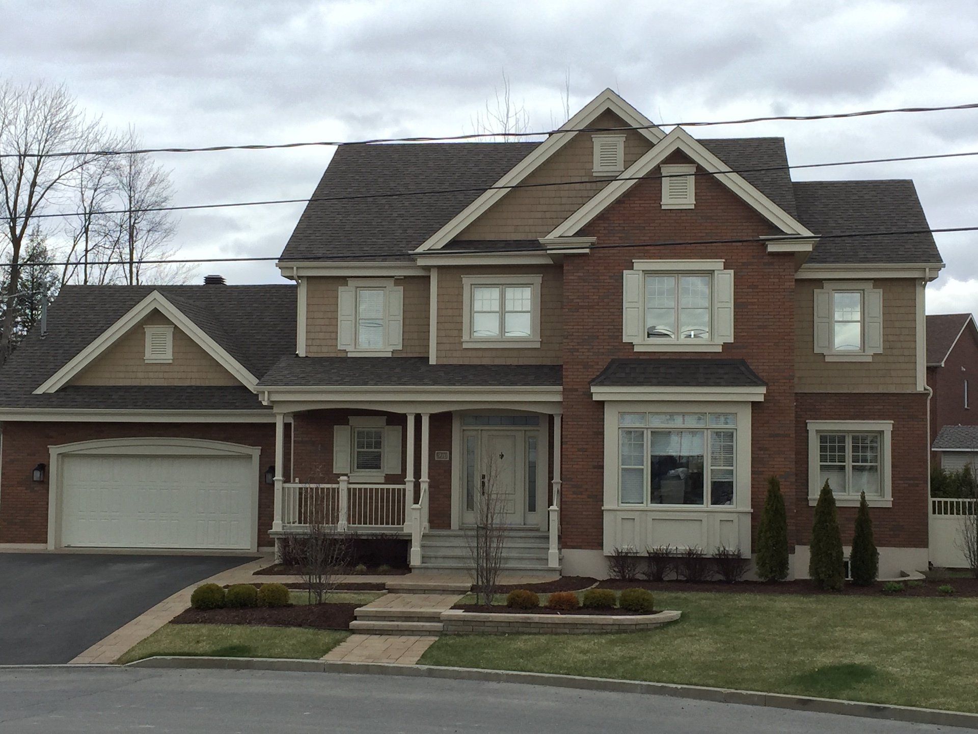 Maison à deux étages avec bardage beige, façade en briques et garage double sous un ciel nuageux.