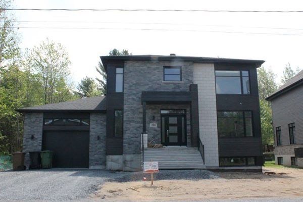 Maison moderne à deux étages avec façade en pierre grise et bardage foncé. Grandes fenêtres, perron et garage attenant.