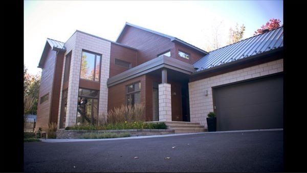 Maison moderne avec bardage brun, éléments en pierre claire et porte de garage grise. Allée asphaltée menant à l'entrée.