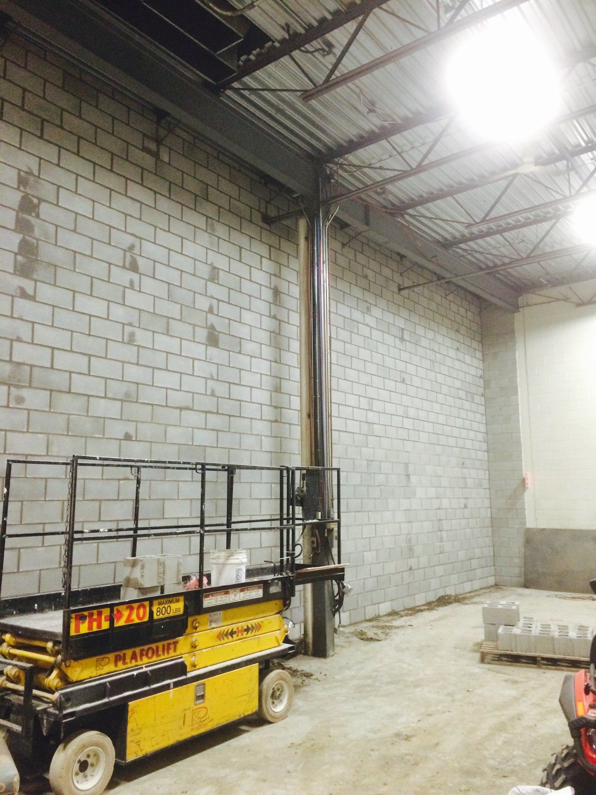 Une nacelle élévatrice jaune est garée à côté d'un mur en parpaings dans un espace industriel inachevé.