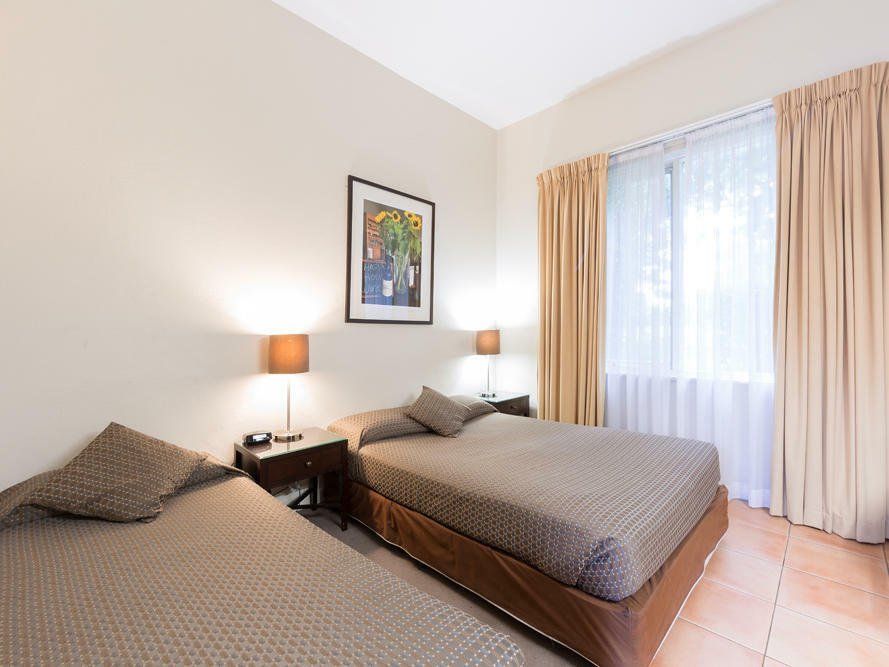 Two twin beds in a bright hotel room with a bedside lamp, framed art, and sheer curtains