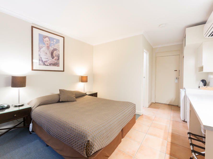 Bedroom with a double bed, bedside lamps, framed art, and a hallway leading to a kitchenette.