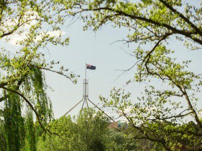 Australian flag atop a tall spire, framed by green tree branches against a pale sky