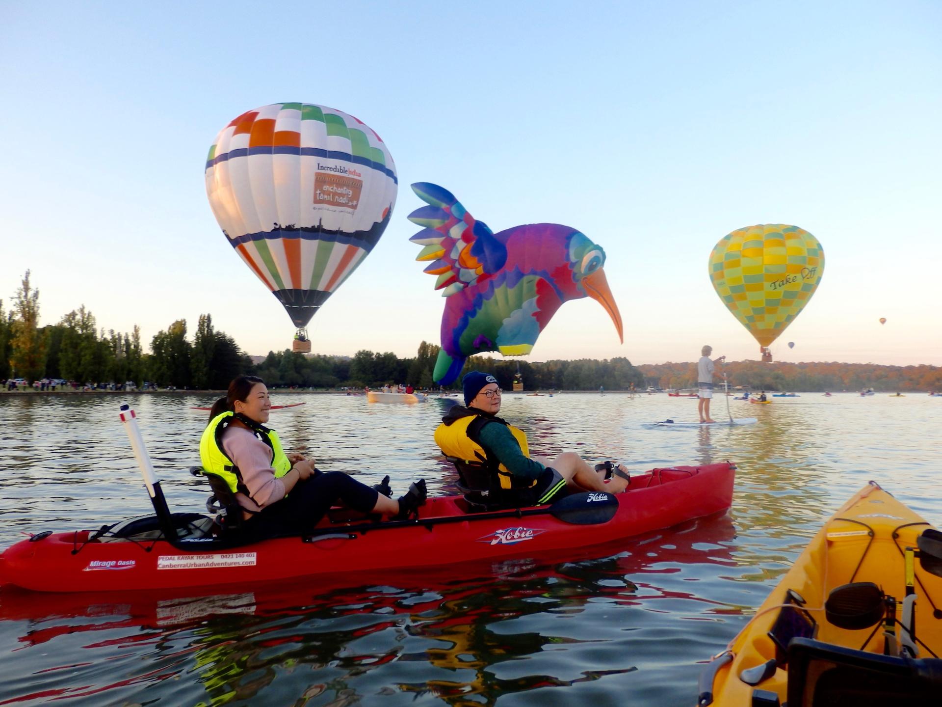 People kayaking on a lake at sunset with colorful hot air balloons overhead