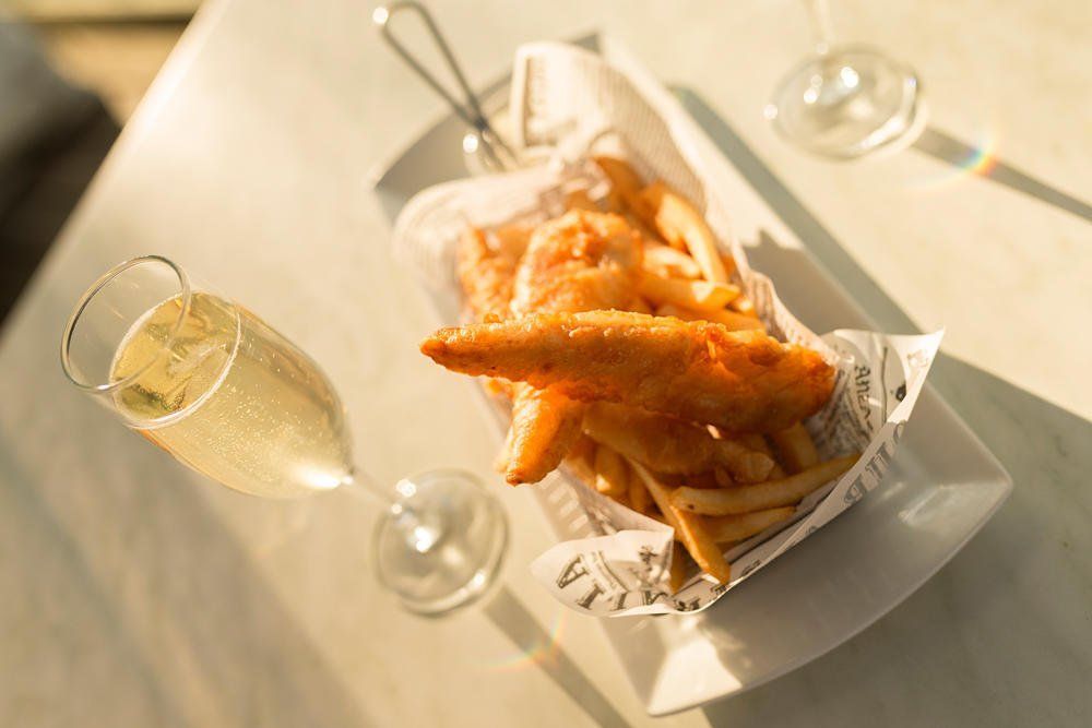 Golden fries in a paper-lined basket beside a glass of white wine on a sunlit table