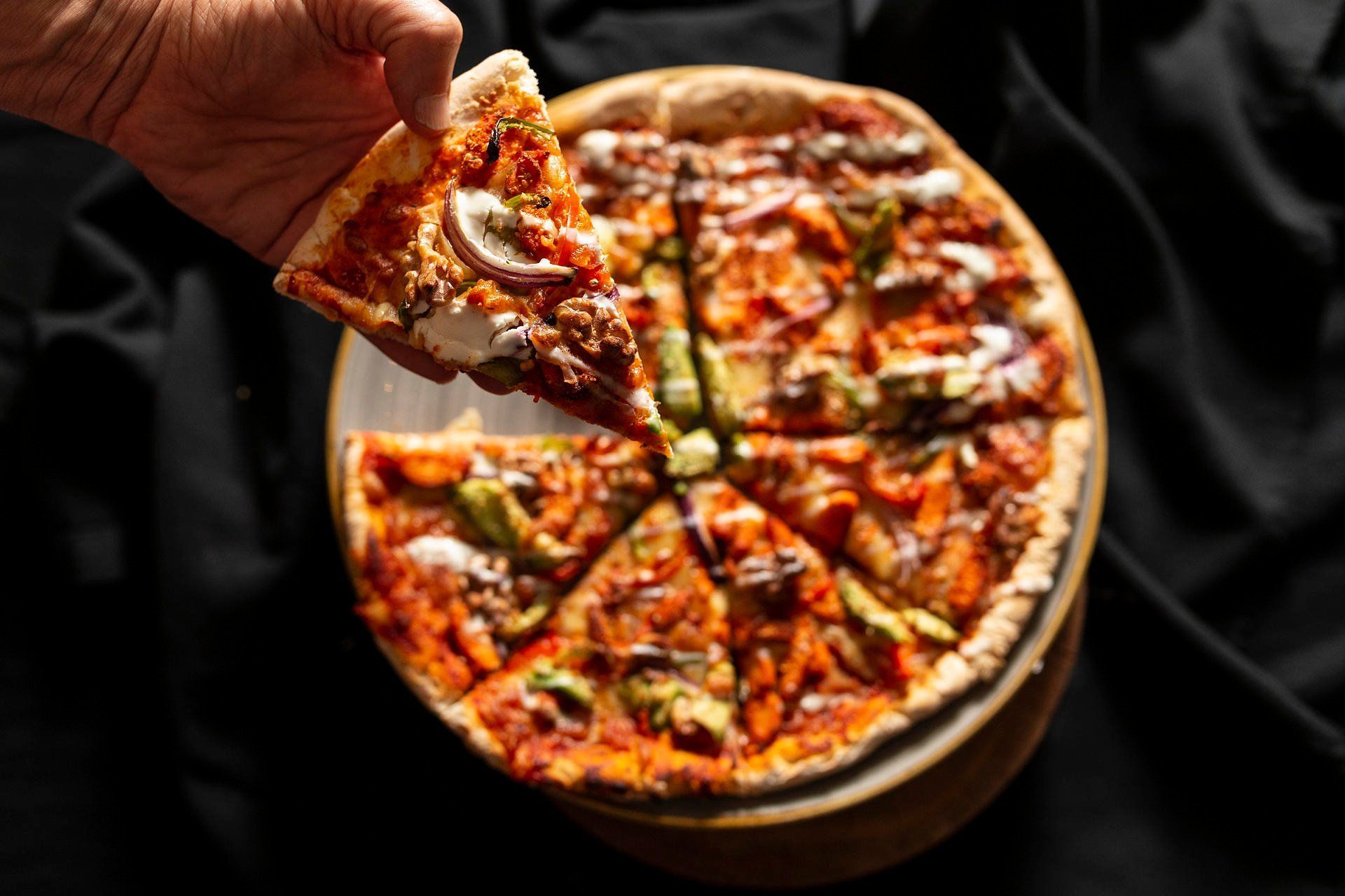 Hand holding a slice above a hot cheese pizza on a dark background