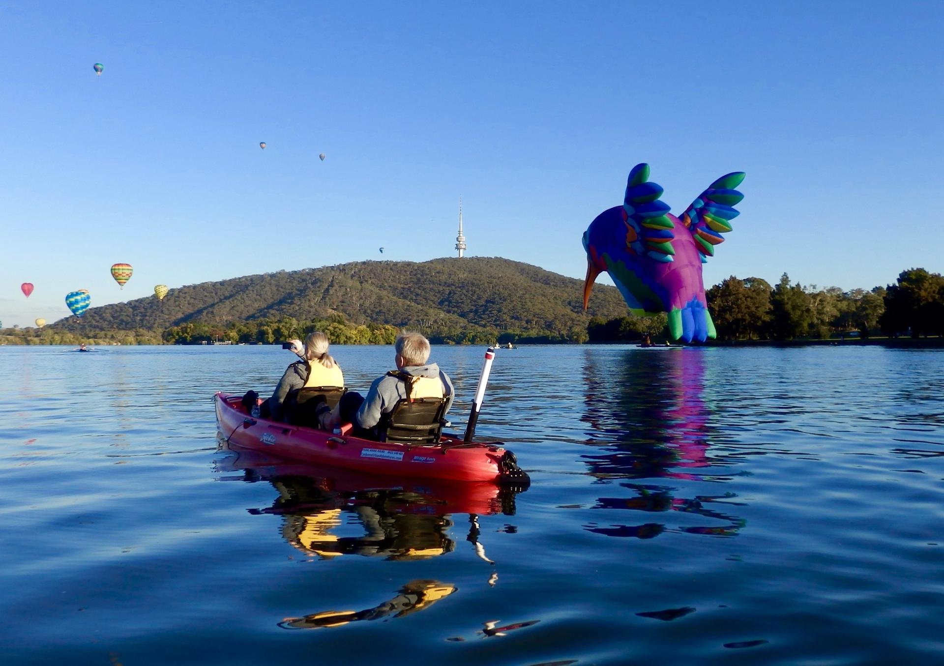 Two people in a red kayak on calm water near a giant colorful bird sculpture at a lakeside festival