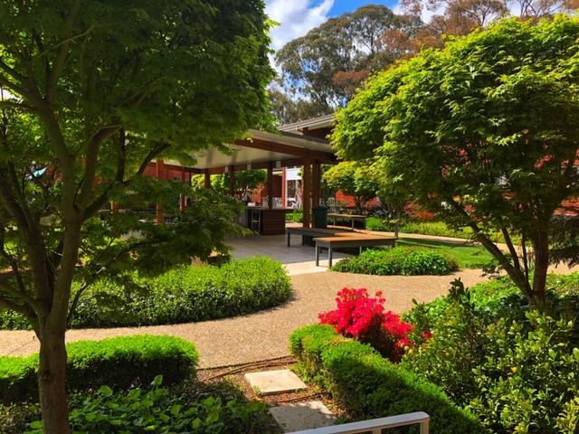 Lush garden path leading to a shaded patio with tables, surrounded by green trees and bright pink flowers