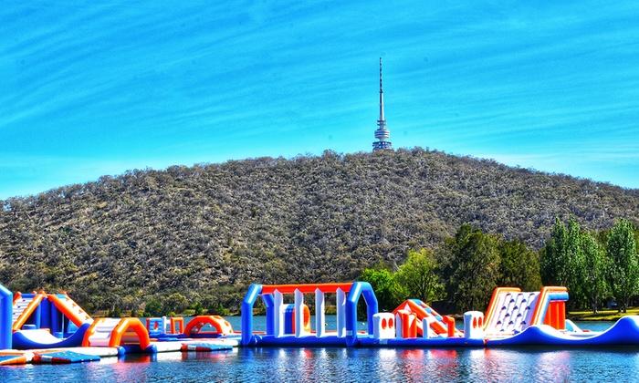 Colorful inflatable water park floats in front of a wooded hill with a tall tower under a bright blue sky.