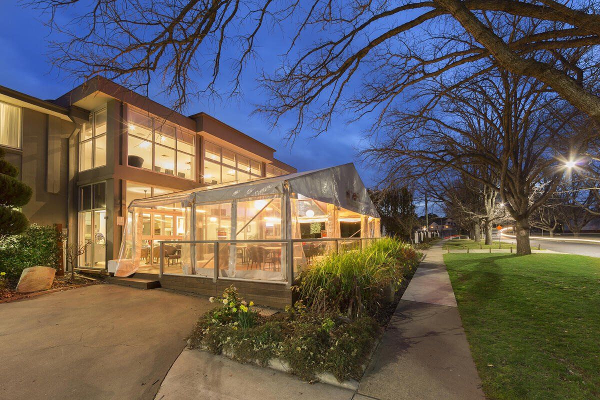 Modern house with lit windows and glass patio at dusk, beside a sidewalk and green lawn