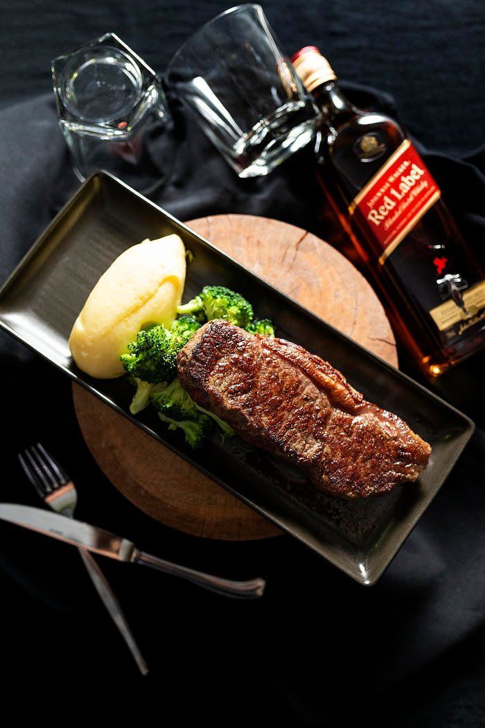 Grilled steak with mashed potatoes and broccoli, served on a black tray beside a bottle of whiskey and a glass.