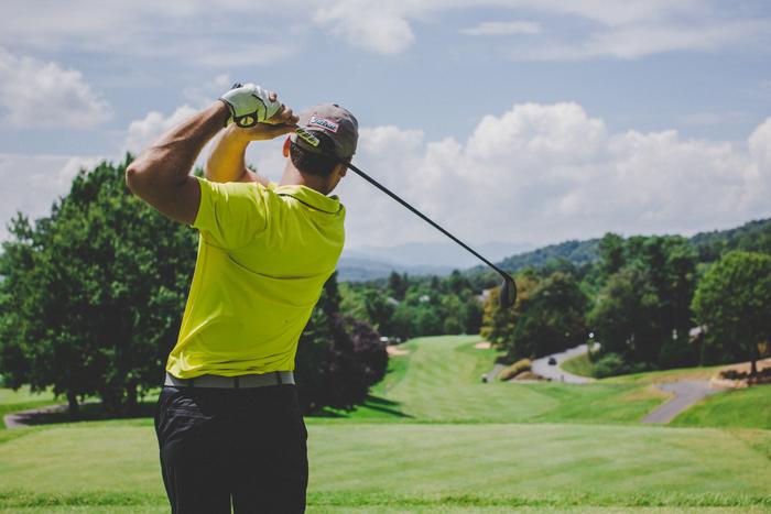 Golfer in a bright yellow shirt swinging a club on a green course with trees and hills in the background
