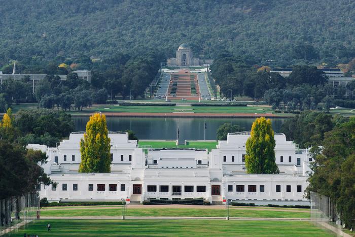 A white temple-like building centered in a green park, with a long staircase and forested hills behind it