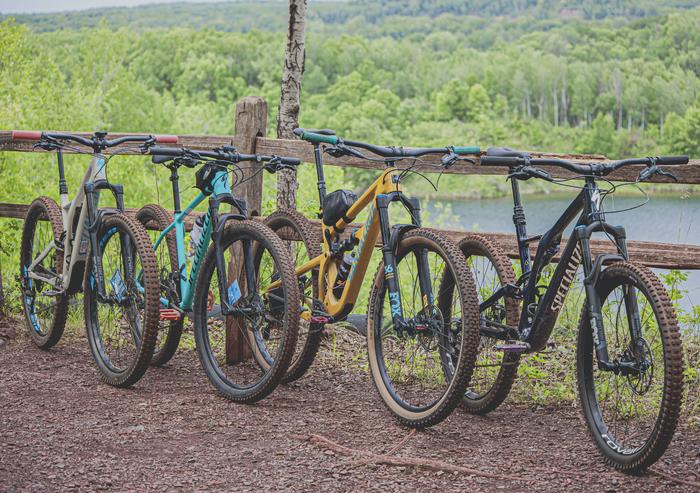 Four muddy mountain bikes lined up by a wooden fence overlooking a green valley