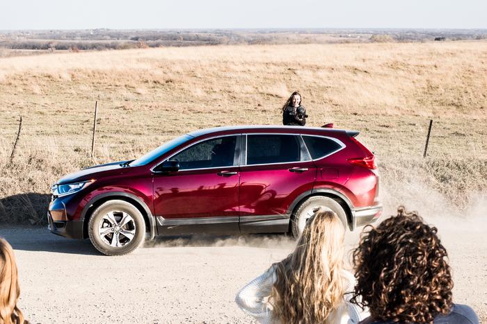 Red SUV driving on a dusty dirt road beside a dry field, with spectators in the foreground.