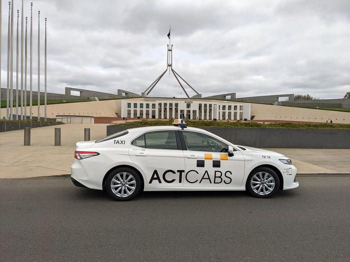 White ACTCABS taxi parked before a large government building with flagpoles and a cloudy sky