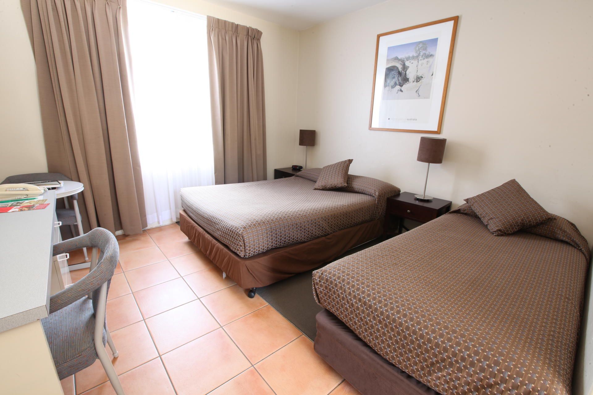 Small bedroom with two brown beds, beige curtains, terracotta tile floor, and framed wall art.