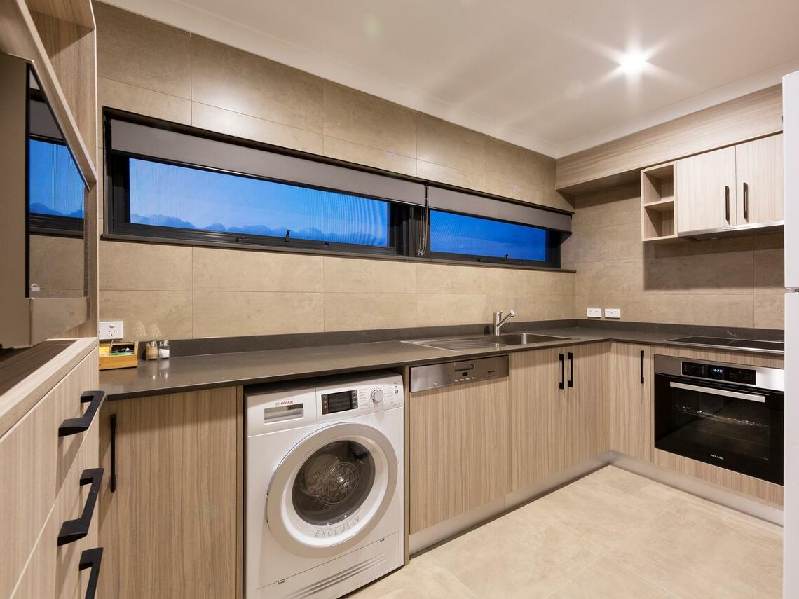 Modern beige kitchen with wood cabinets, black countertops, a window above the sink, and a washing machine under the counter.