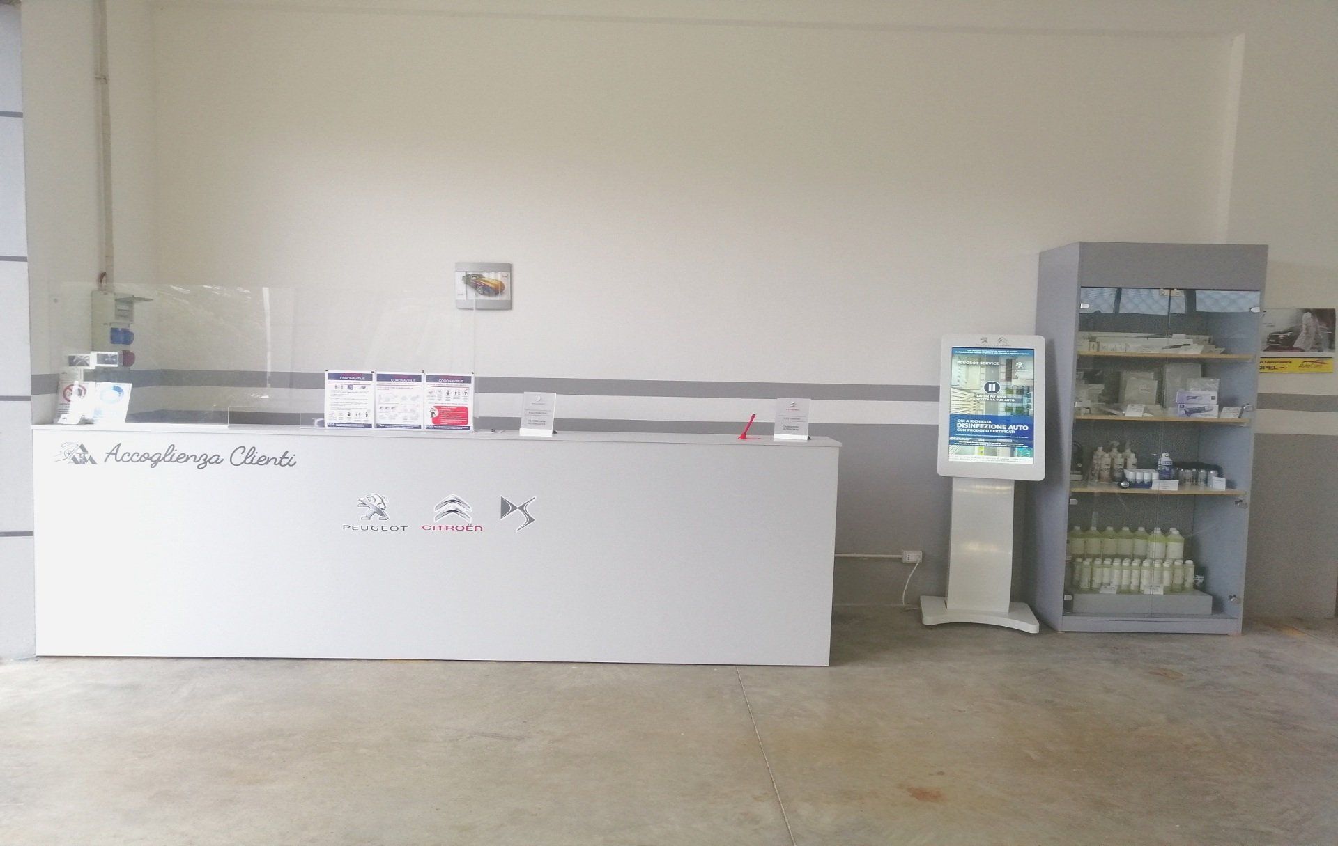 Un bancone bianco, prodotti esposti in un negozio o in un ufficio, uno schermo digitale e una scaffalatura grigia.
