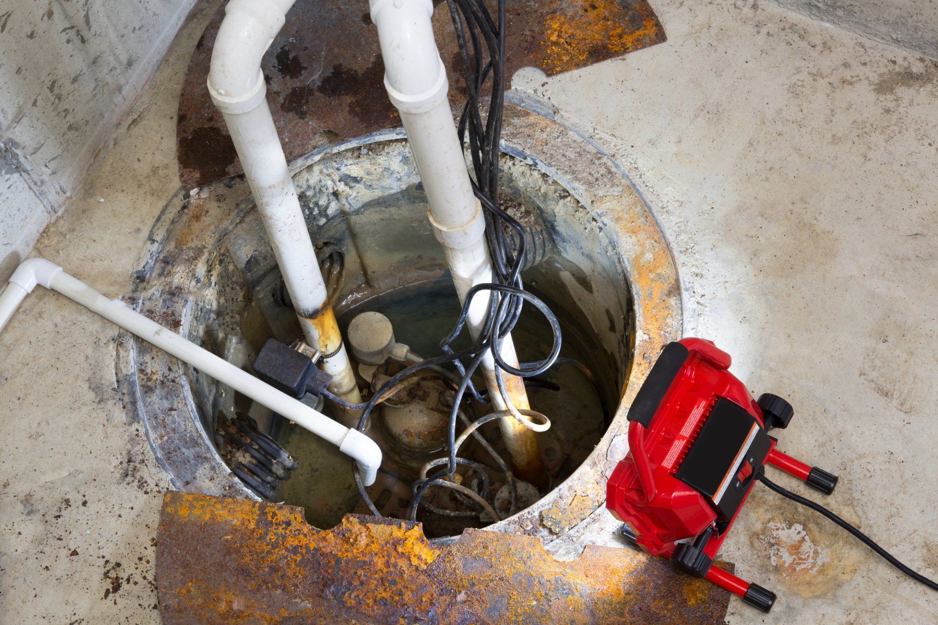 Sump pit with pipes, a pump, and a work light. Concrete walls are visible.