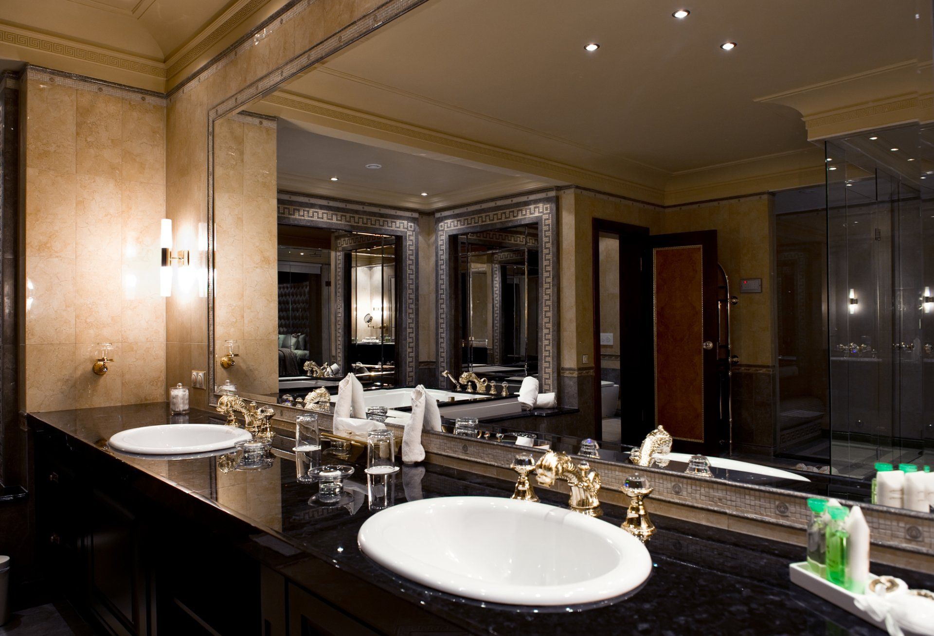Luxurious bathroom with black countertop, gold fixtures, and two white sinks. Mirrored walls and tub visible.