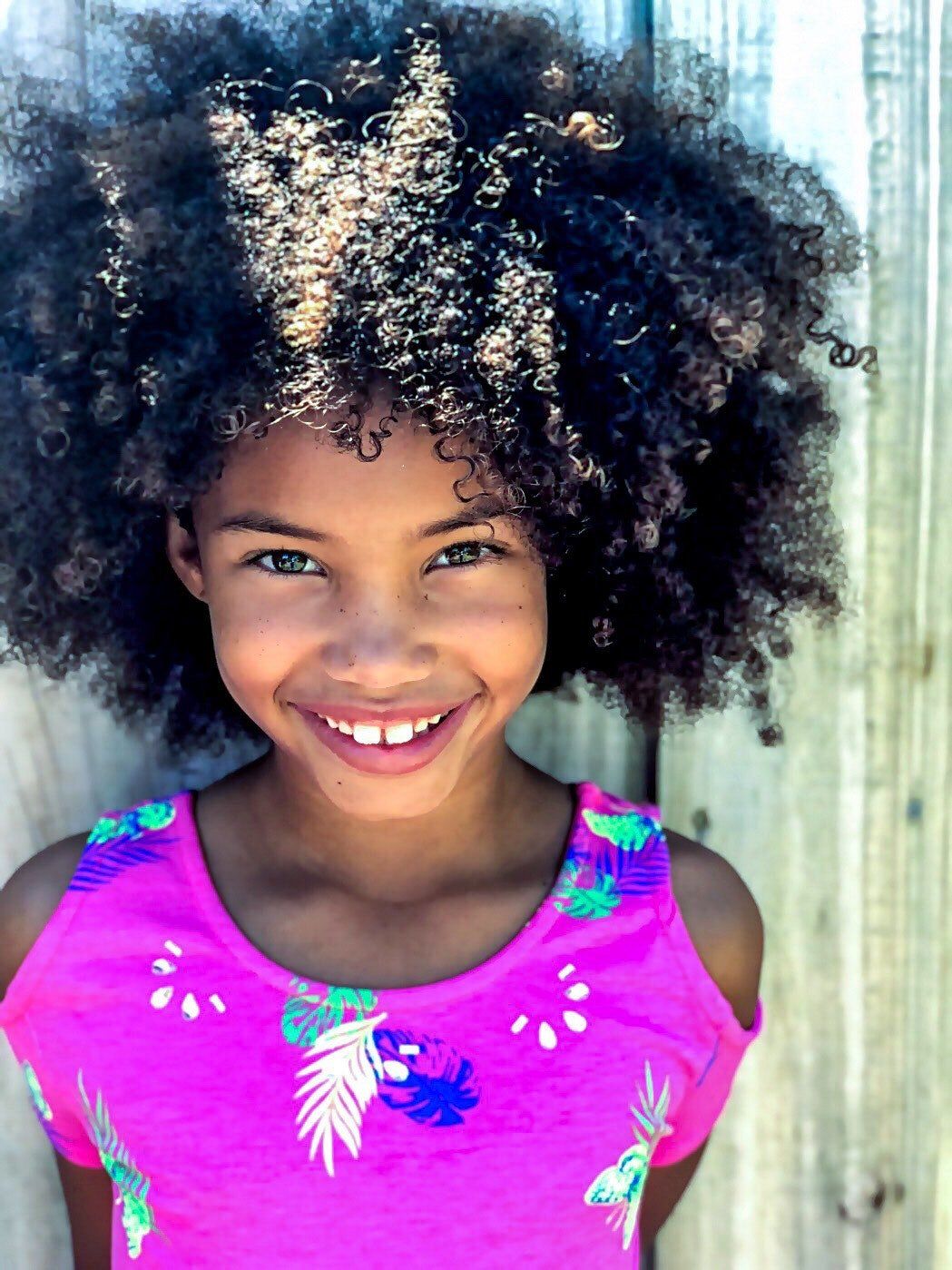 A young girl with curly hair is wearing a pink shirt and smiling.
