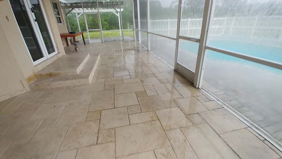 Wet stone patio beside a covered pool, with glass walls and a grassy yard beyond.