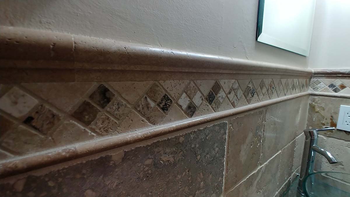 Close-up of a tiled bathroom or kitchen wall with beige diamond trim and a faucet at right.